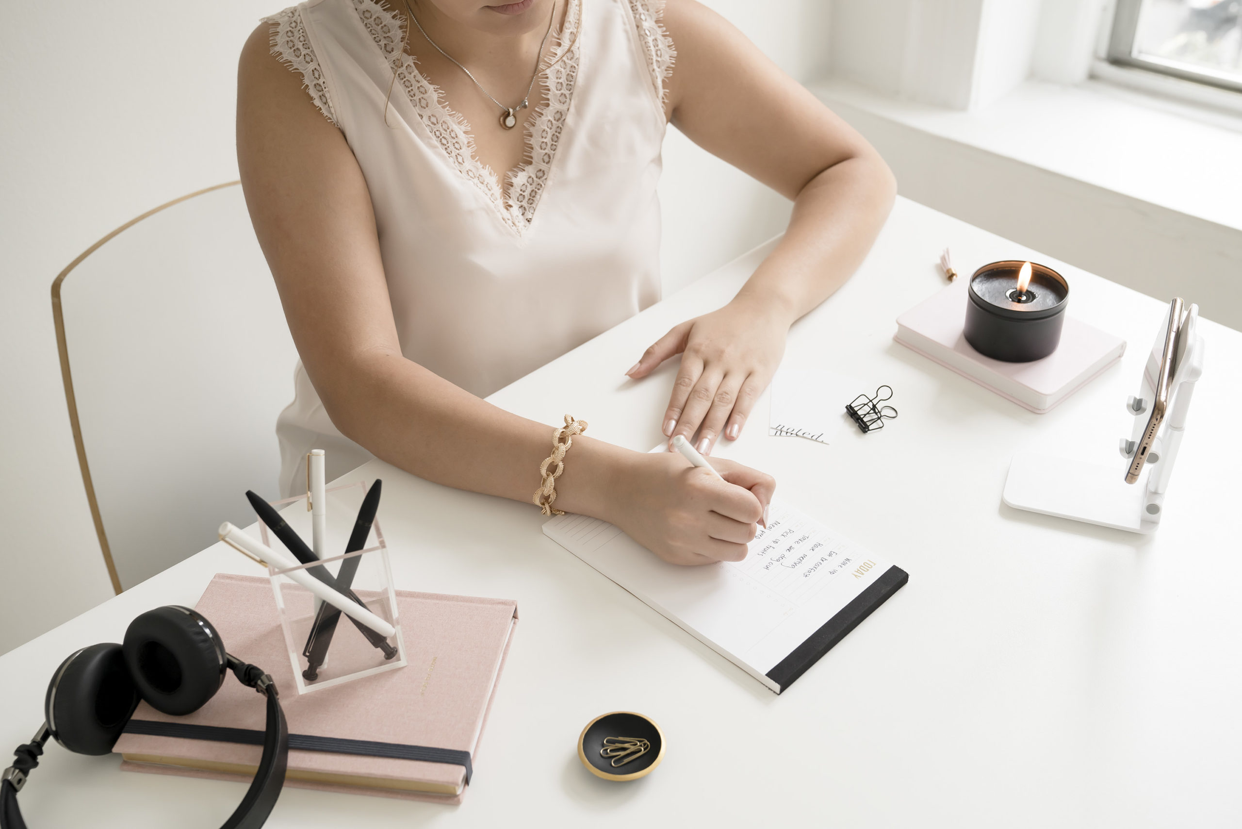 A woman in white sits at a white desk with pink and black office accessories writing a to-do list for today terminating the underperformer