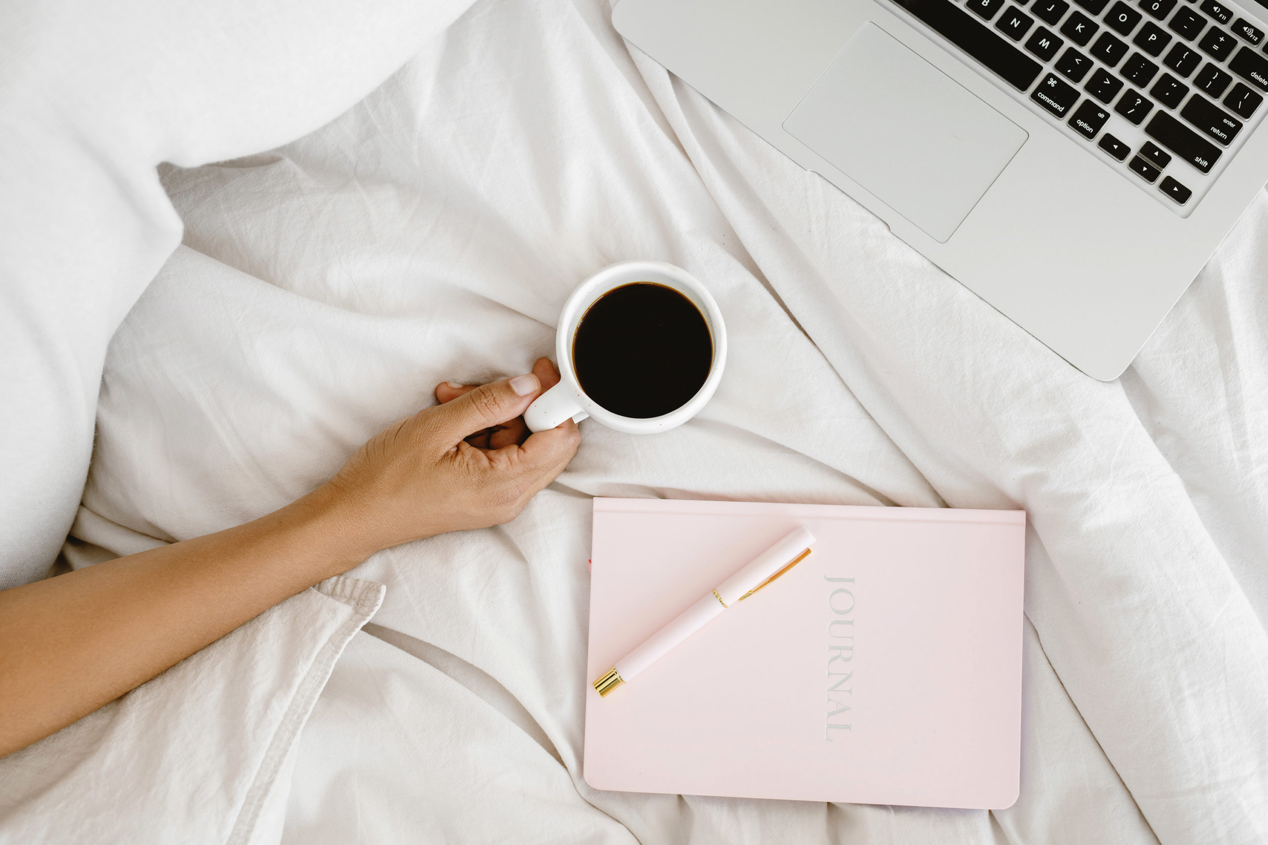 A cozy bed with white linen and a hand holding a coffee along side a pink notebook and computer at the edge of the picture!