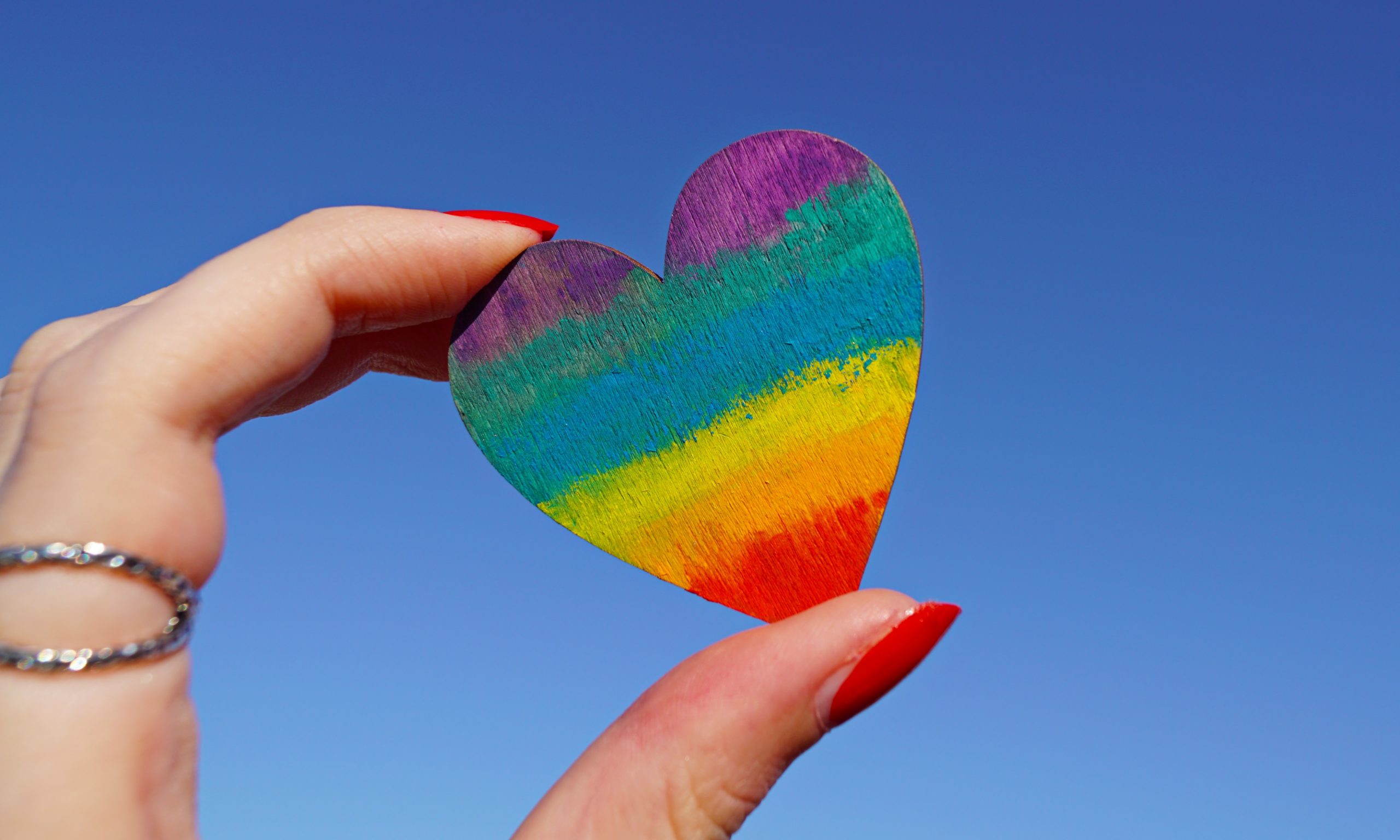 hand with red painted nails holding a rainbow heart against blue sky