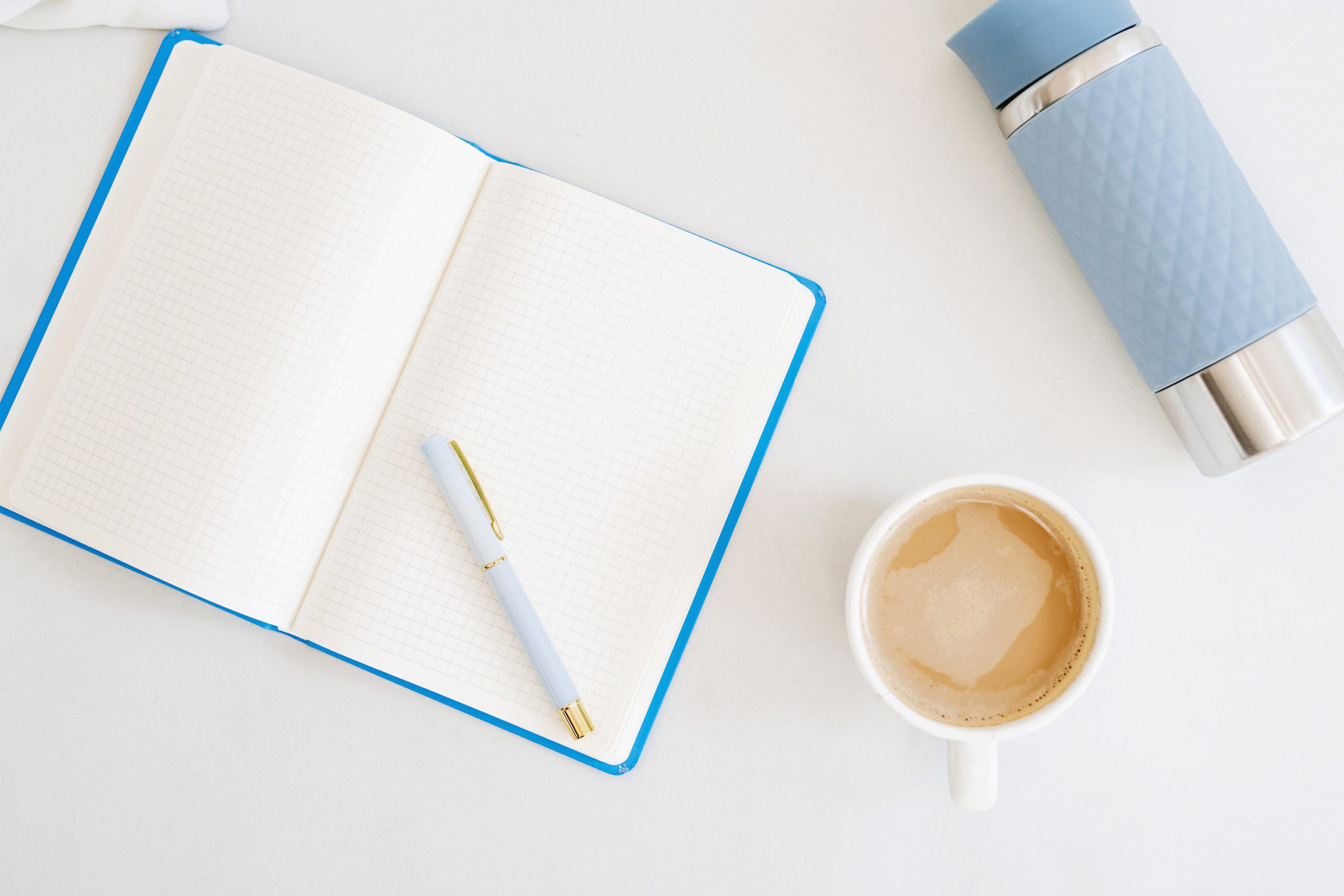 A blue notebook lies open on a white table with a coffee and a blue thermos lies next to it