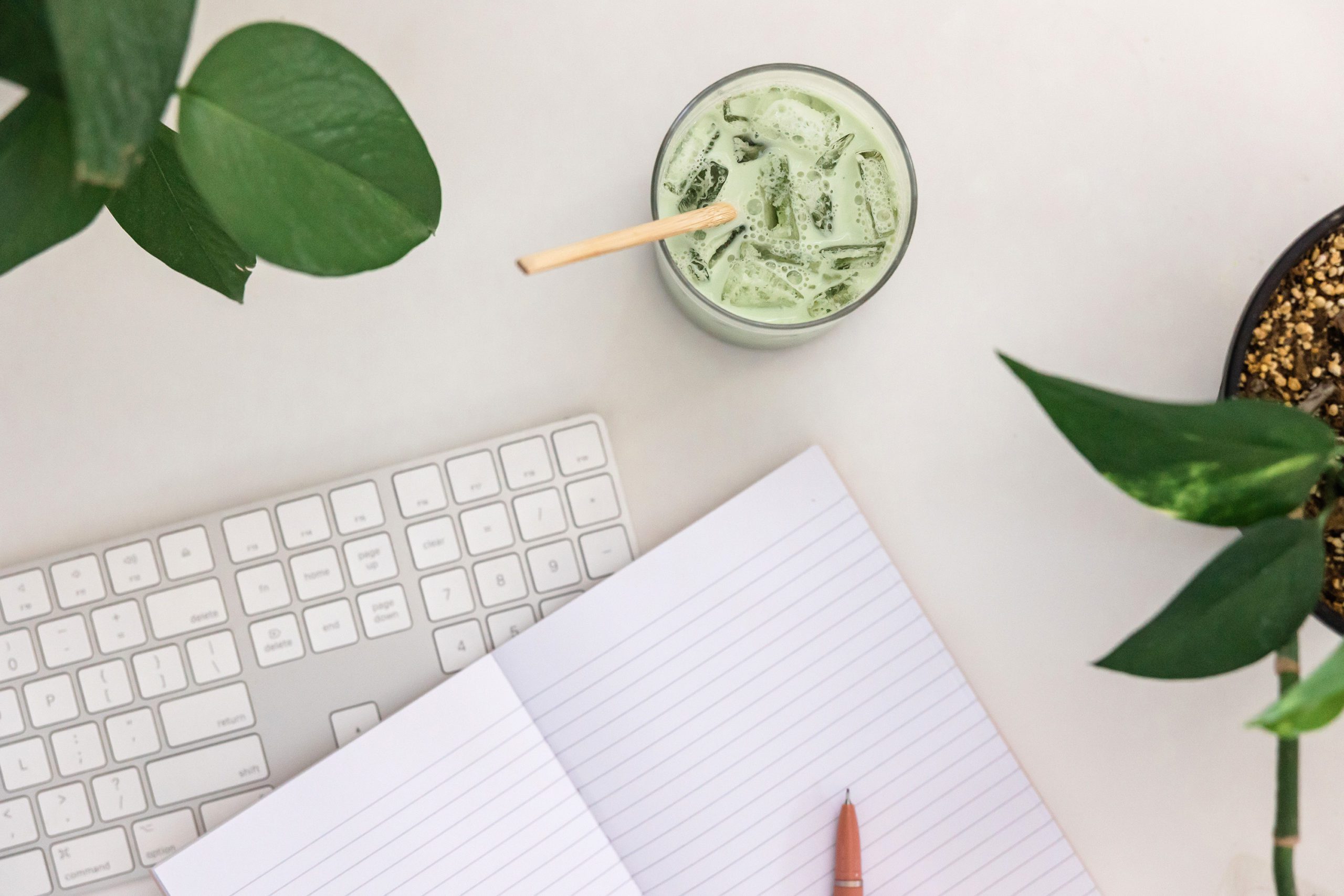 A white workspace with a keyboard, notebook, matcha latte and plants