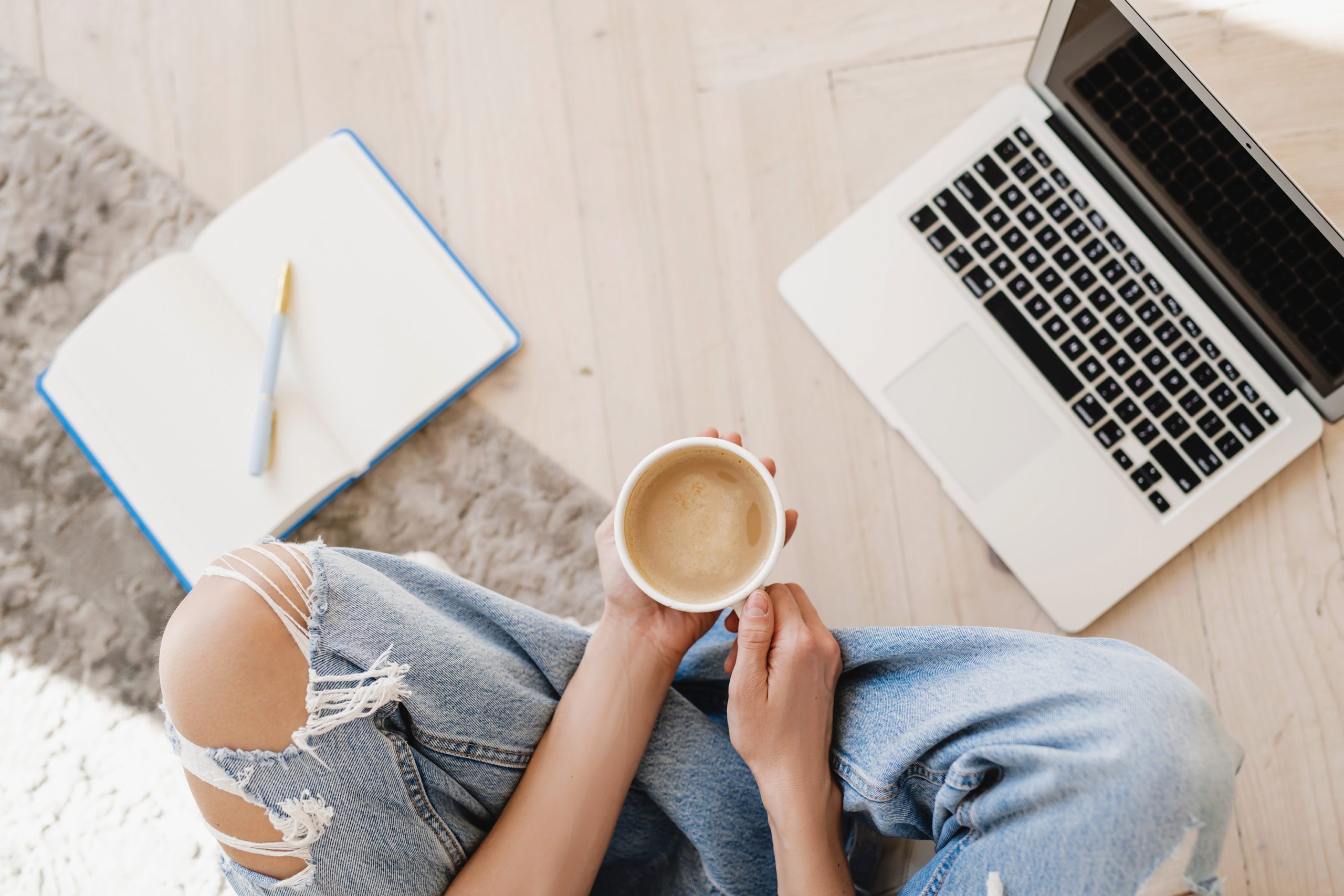 A woman in ripped jeans holding coffee sitting cross legged on the floor with a blue notebook and laptop